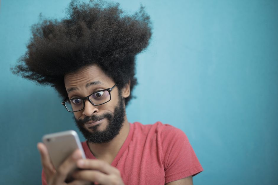 Um homem surpreso com cabelo afro e óculos olha para seu smartphone contra um fundo azul.