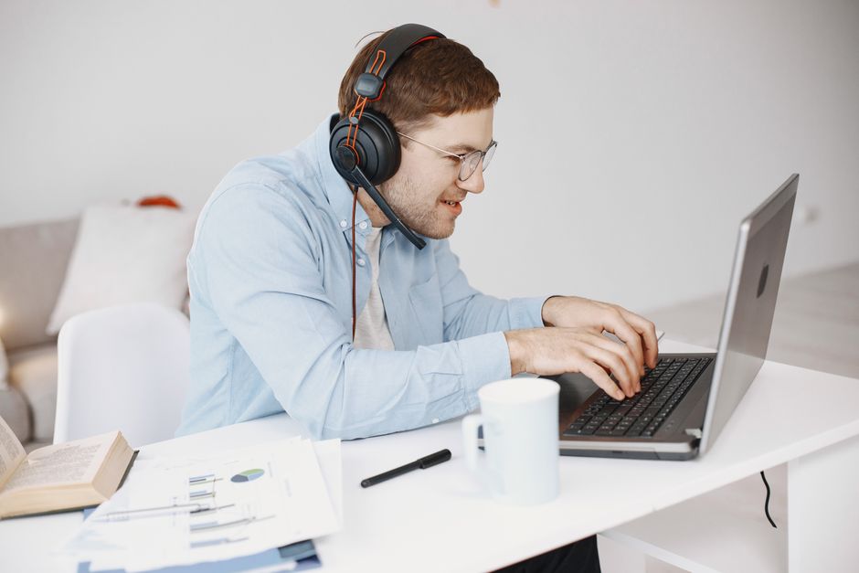 Homem adulto usando laptop com fones de ouvido para trabalho remoto no escritório em casa.
